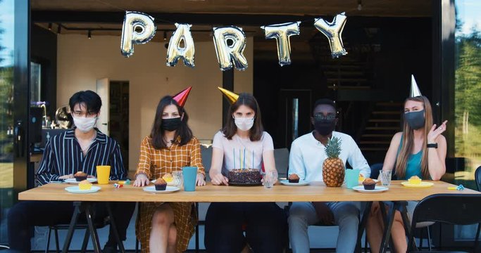 Weird party celebration after COVID-19. Group of sad upset multiethnic friends in masks sitting at table slow motion.
