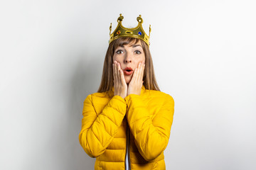 Young woman in a yellow jacket and a crown on her head with a surprised face on a light background. Emotion laughter, surprise, kiss. The concept of a queen, luck, the best, win, victory. Banner