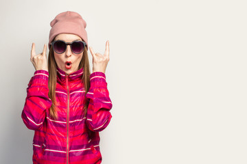 Young woman in a hat and glasses and a pink sports jacket makes a rock and roll goat gesture, we'll catch this party on a white background with empty space. Concept. Face expression. Banner