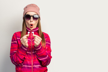 Emotional Young woman in glasses, a hat and a pink sports jacket with a surprised face holds wireless headphones on a white background. Concept modern style, cool music. Face expression. Banner