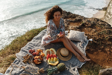 Happy caucasian woman in straw hat with curly hair enjoy picnic by the sea, sit on sea cliff and eat fruits - Powered by Adobe