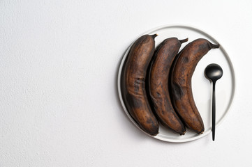 Waste food concept. Ugly food. Stale dark bananas in a white plate and a black spoon on a white background. Top view, copy space.