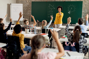 Happy black teacher pointing at his elementary students who are raising hands to answer the...