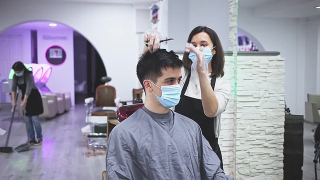 Young woman stylist wearing face mask and gloves working in hairsalon doing haircut and hair styling for male client, new normal due to coronavirus outbreak