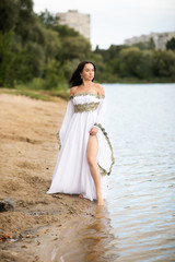 Young european woman with brown hair dressed a long white wedding dress stays near the river