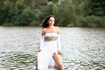 Young european woman with brown hair dressed a long white wedding dress stays in the river