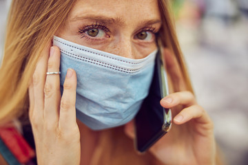 Red haired pretty masked girl with freckles talking on smartphone close up