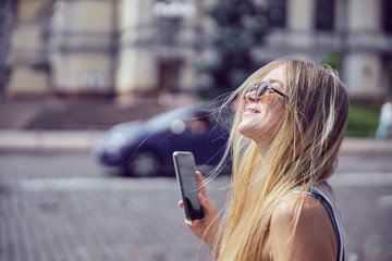 Cheerful pretty girl with long hair in sunglasses holding a smartphone outdoors