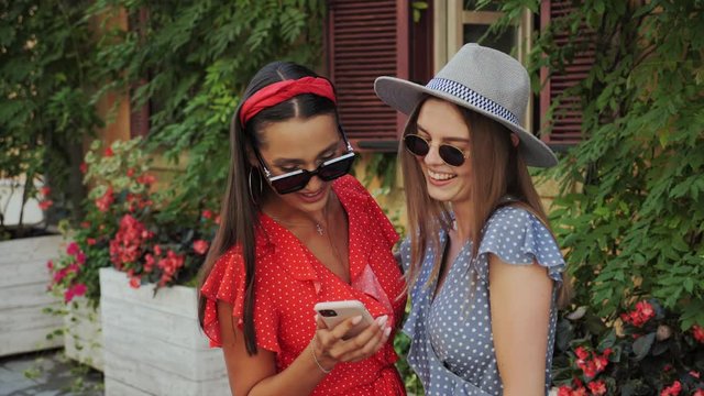 Two best cheerful sisters in a sunglasses and hat have fun, laugh, take selfies and look to the pictures on the background of a beautiful house with flowers in the city.