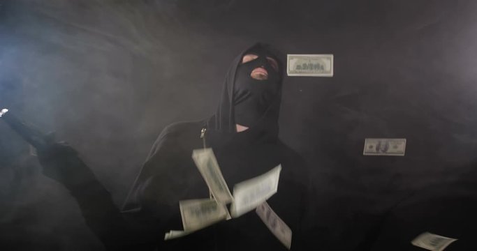 Robber  in black clothes, mask and gloves with a gun dancing, posing and taking aim, on black studio background with smoke and money falling on him. Security system concept. 