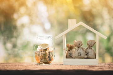 Money bags put in the wooden home model and bottle with gold coin inside on sunlight in the public park, Saving money or loan for business investment real estate or buy new home for a family concept.