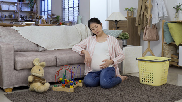 Full Length Pregnant Housewife Collecting Toys In Living Room Is Taking A Breaking, Stroking Her Tummy. Tired Looking Asian Woman With Swollen Belly Sitting On Carpet Is Picking Up After Her Child