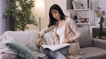 attractive asian woman in casual attire is spending time with herself reading in living room....