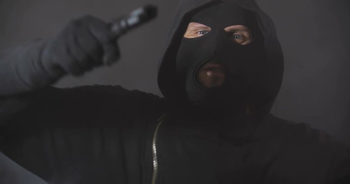Close up robber in black clothes, mask and gloves with a gun talking and shouting aggressive to camera on black studio background with smoke. Security system concept.  4K 50 fps slow motion