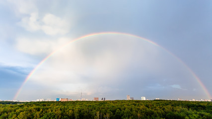 rainbow in blue cloudy sky over green forest and residential district in city in sunny summer evening