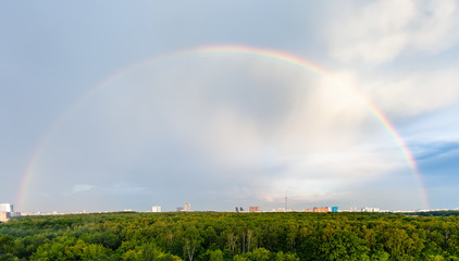 panoramic view of rainbow in blue cloudy sky over green forest and residential district in city in sunny summer evening