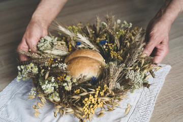 Woman's hands are picking up Polish harvest wreath for harvest festival from wooden table. The wreath is made of wheat, oat, Flaxes, wild herbs and wildflowers, yarrow, tansy, cornflower and chamomile