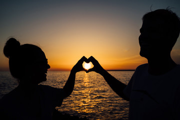 Couple making a heart symbol at sunset with blue and orange sky. - Powered by Adobe