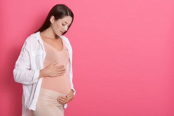 Beautiful pregnant woman hugging her tummy against pink background, looking at her belly with love, copy space for advertisement or promotional text text.