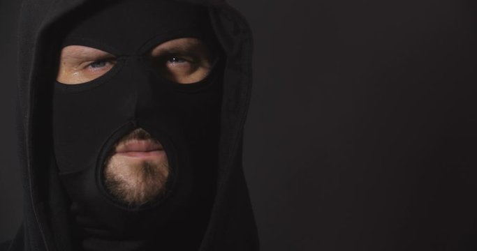 Close up portrait of robber in black clothes and mask looking aggressive to camera on black studio background with smoke. Security system concept.  4K 50 fps slow motion
