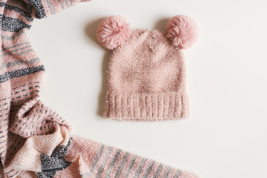 Handmade knitted hat with two pompoms and warm scarf of pink color on white background, top view. Autumn and winter season clothing