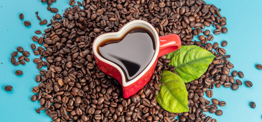Red cup in the shape of a heart with freshly brewed black coffee and coffee beans scattered on a blue background