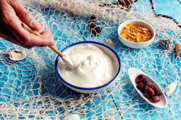 Greek yogurt, cereals and red berries on a blue background next to a fishing net and seashells.