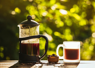 French press coffee and cup on table with scattered coffee beans in garden on sunny bright morning day. Reason to wake up concept. Bokeh green background with room for text.