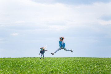 little girl child and mother woman run and jump, green grass in the field, sunny spring weather, smile and joy of the child, blue sky with clouds