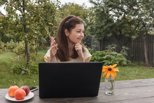 Young woman remotely working on a laptop sitting at a wooden table on a background of green garden and looking at the side. online training. online shopping. happy mother. online break