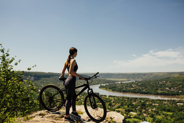 Fototapeta na wymiar Woman cyclist standing with bicycle on rock on mountain. Young athlete biker with mountain bike on top of the hill observing the river and town view under blue sky.
