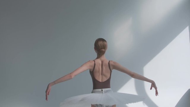 Fragile ballerina on a white background standing with her back performs smooth movements with her hands. Graceful rehearsal of the part of the white swan in a spacious hall. Close up. Slow motion.
