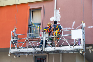 Two plasterers painters performing painting the facade of the house staying in the building cradle....