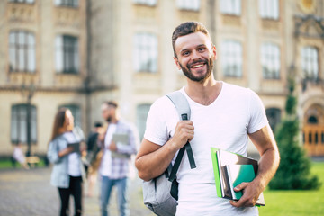 Young handsome bearded student guy in casual clothes with backpack and books in hands is walking and posing on a university  buildings background