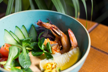 Bowl poke with fried shrimps, cucumber, lemon, tomatoes is on the table in a cafe
