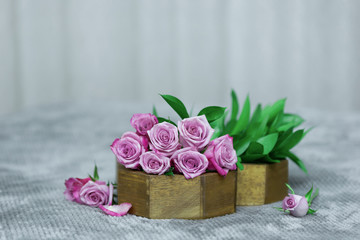 Purple rose bouquet in brown wooden vase with green leaves on gray background with petals on gray fluffy surface
