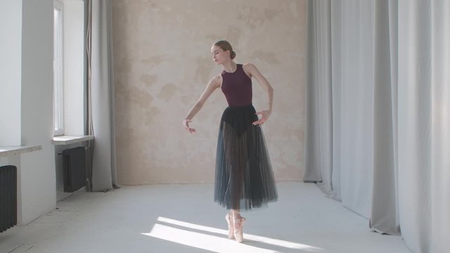Professional performance of a fragile ballerina against the background of large panoramic windows and curtains. Filmed in a loft style studio bathed in bright daylight. Slow motion.