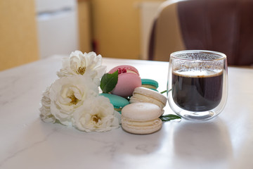 Colourful macaroons are laying on the table along with the coffee in transparent cup and white roses, breakfast time