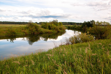 a narrow river with a mirror of blue sky with clouds, with a high Bank on the other side