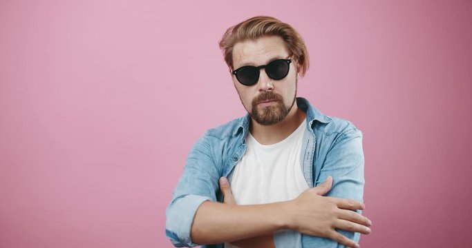 Handsome bearded man with stylish haircut wearing black sunglasses, white t-shirt and denim shirt posing on camera. Isolated over pink studio background.