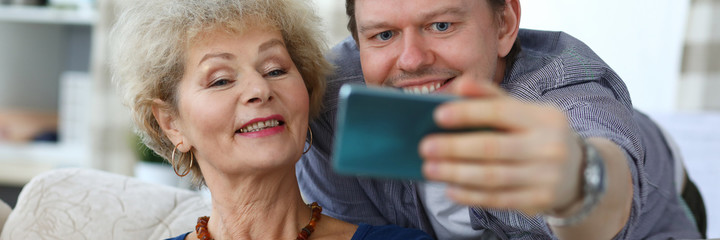 Portrait of cheerful son and mother taking selfie indoors. Happy family resting together at home. Smiling middle-aged man and senior female on sofa. Relationship and leisure concept
