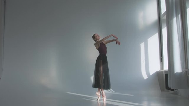 Ballerina in black ballet tutu dress dancing classical pas in class near the window. Alone warming up before performance. White background of a spacious studio bathed in bright daylight. Slow motion.