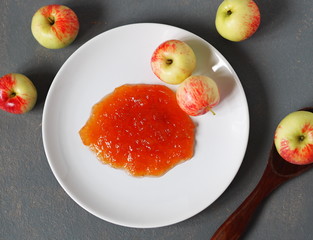 Homemade apple jam in a white plate on a gray background with fresh apples and a wooden spoon.Healthy natural food.