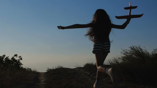 Silhouette rear view of a girl with very  long brown hair , having  fun, running with a red toy airplane in the countryside with clear blue cloudless sky in background. 200 fps slow motion 