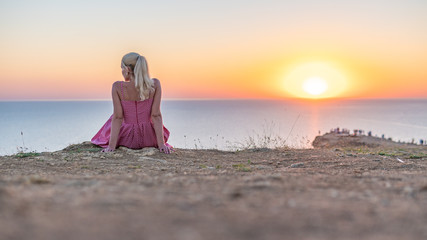 A joyful adult girl with a Caucasian appearance looks at the sunset with her back in the summer in the evening