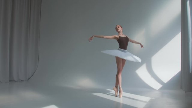 Female dancer does ballet exercises in stage dress with open back. Rehearses dance moves in the spacious and brightly lit studio.