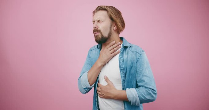 Bearded man in casual clothing sneezing and coughing at studio with pink background. Mature male suffering from sore throat. Concept of illness.