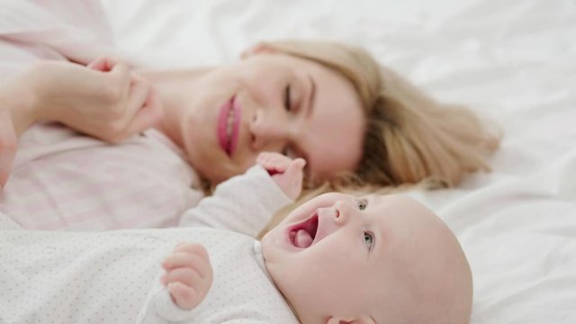 Young mother blonde in pajamas hugs and takes care of a little baby girl in sliders. A mother and child lie on a bed with white linen against a large window with a curtain and a white brick wall.