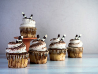 Tiramisu cupcakes lined up with white frosting piled high and sprinkled with espresso powder and a white and dark chocolate stick with a raised cupcake behind them.  Delicious dessert.