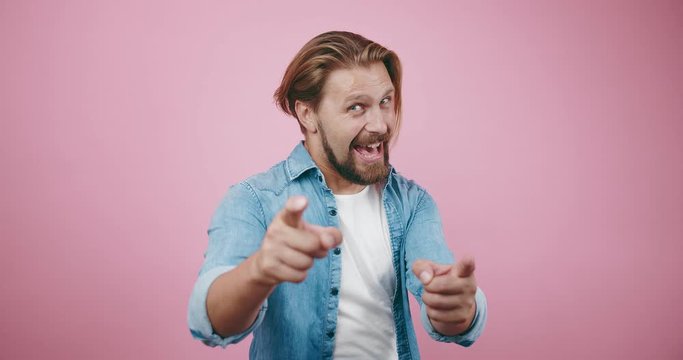 Emotional man with trendy haircut pointing with forefingers on camera over pink background. Bearded male in denim shirt gesturing in studio.